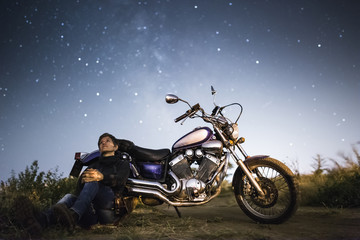 Young beautiful biker woman with motorcycle under stars of  home galaxy Miky Way. Female biker on field at night