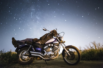 Young beautiful biker woman with motorcycle under stars of  home galaxy Miky Way. Female biker on field at night