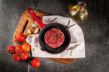 Fototapeta na wymiar Raw organic beef meat burgers cutlets with spices, thyme, tomatoes, olive oil in frying pan on black background, copy space top view