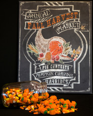 A glass jar has tumbled and spilled all of its candy corn on a table at a fall harvest festival.