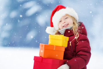 Adorable little girl wearing Santa hat holding a pile of Christmas gifts on beautiful winter day