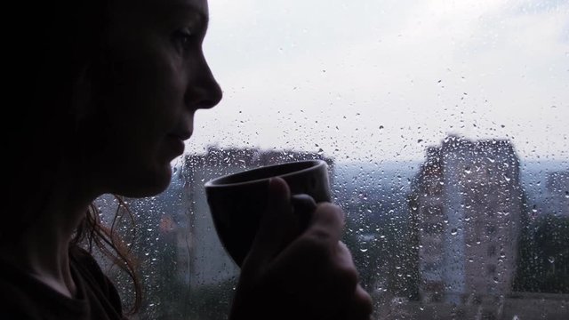 A girl is drinking coffee by the window. Silhouette of a girl by the window with rain.