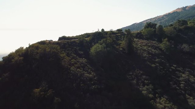 Aerial flying over forest covered hills revealing a car at the edge of a cliff by the ocean. Winding road through the mountains, and fog covered ocean in California.