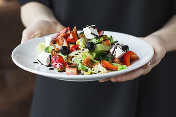 The young female waiter, holding in her hands plate with the Greek salad