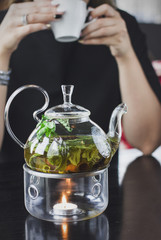 The Glass teapot with herbal tea on the stand with candle on the background of young woman with a mug of tea