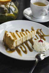 Cheesecake drizzled with caramel on the white plate, kettle with herbal tea on black table