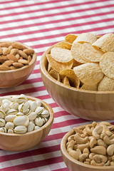 Bowls of potato chips, almonds, pistachios, cashews on red checkered tablecloth