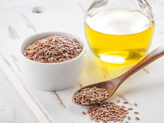 Brown flax seeds in spoon and flaxseed oil in glass bottle on white wooden background. Flax oil is...