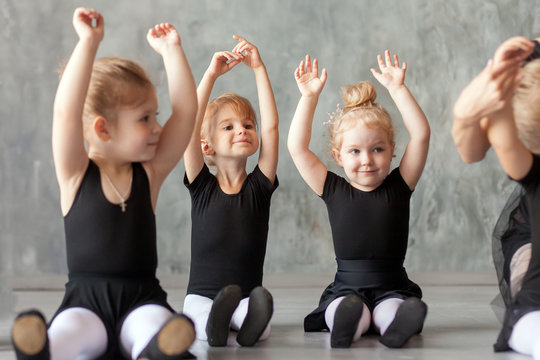 little girls ballerina in black dresses, belk tights and pointe shoes sit on the floor and hold hands over their head for warm-up before teaching ballet in a dancing dark studio