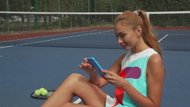 Girl sitting on a tennis court and holding smartphone. She looks in front of her, then lowers her gaze to phone. Racket, balls ant tennis net are behind her