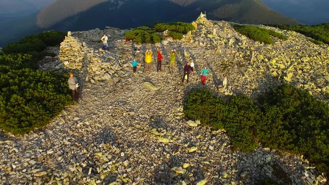 Flight over the company of friends who stand on top of the mountain, during the sunset, waving their hands, having fun, enjoying a successful journey. Epic adventure in the mountains.