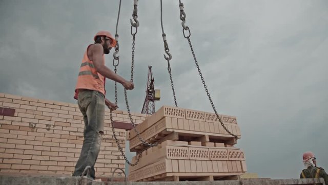 The epic builder works against the gray cloudy sky and clings bricks to the crane hooks. Construction site: building a brick house. Brutal male builders at work. Civil engineering. Hard work for men.