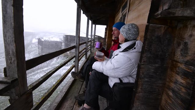 Couple of travelers is drinking coffee on the veranda of a abandoned wooden hut during a storm in the mountains and looking at the snowfall. Epic travel in a mountains of Georgia.
