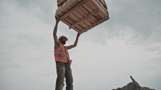 An epic builder works against a gray cloudy sky and gives a command to a crane operator. Construction site: building a brick house. Brutal male builders at work. Civil engineering.
