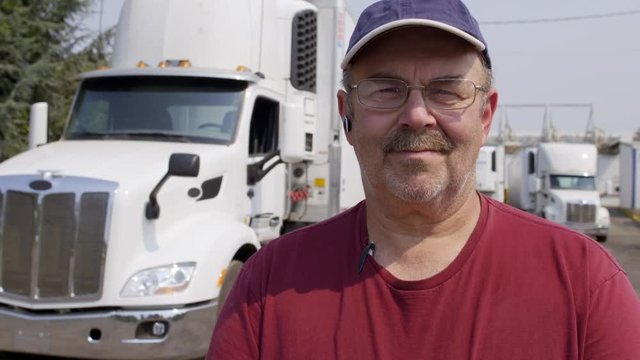 Close up portrait of driver wearing burgundy T-shirt, cap and glasses, looking at viewer and standing in front of truck in haulage yard. Slow motion dolly from right, 4K recorded at 60fps