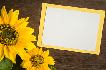 White blank greeting card with beautiful yellow sunflowers on the brown wooden table. Empty place for a text. Top view.