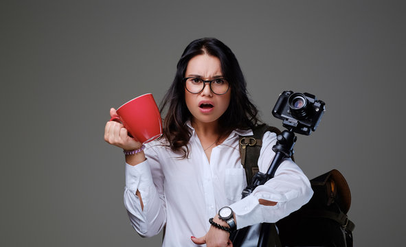 Female holds digital photo camera and a red coffee cup over grey background.