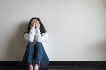 Stressful depressed woman sitting sadly with mental health illness, headache, migraine and emotional anxiety or panic disorder on the floor in home living room with white wall  