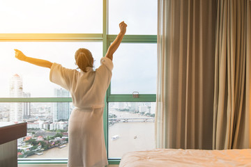 Easy lifestyle Asian woman waking up in morning taking some rest relaxing in hotel room or home bedroom with blur city background view of office towers and residential buildings in business downtown © Chinnapong
