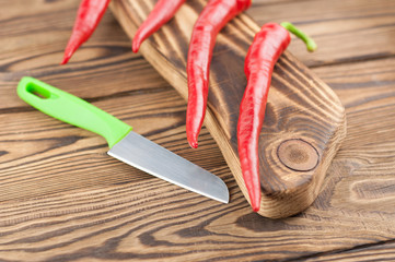 Metal knife with green plastic handle beside row of wholes red fresh ripe chili pepper on cutting...