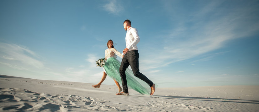 Bride and groom hold hands, smile and walk barefoot in desert. Widescreen image and bottom view.