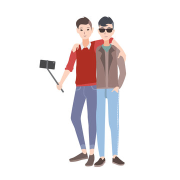 Two young men dressed in stylish clothing standing together, smiling and making selfie photo using monopod with smartphone