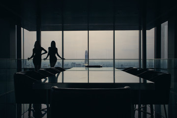 Two woman silhouette standing behind meeting table opposite panoramic window with modern city line under sunset