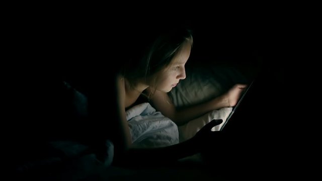 Young, happy caucasian girl watching movie on tablet computer while lying on bed at night