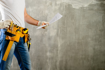 unidentified handyman with hand on waist and tool belt with construction tools holding the project plane against grey background, copyspace. DIY tools and manual work concept