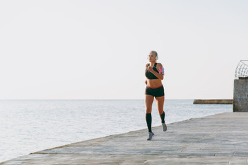 Young beautiful athletic girl with long blond hair in headphones listening to music and running at sunrise over the sea