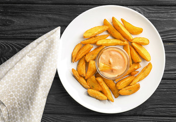 Plate with delicious baked potato wedges and sauce on wooden background