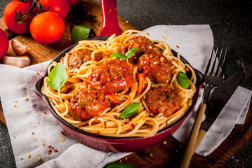 Spaghetti pasta with meatballs, basil tomato sauce in red cast iron pan, on black stone table with cutting board copy space