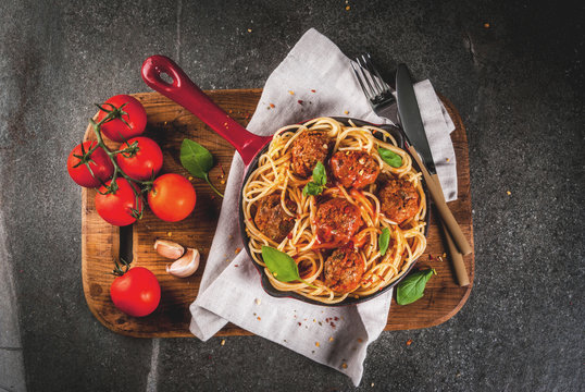 Spaghetti pasta with meatballs, basil tomato sauce in red cast iron pan, on black stone table with cutting board copy space top view