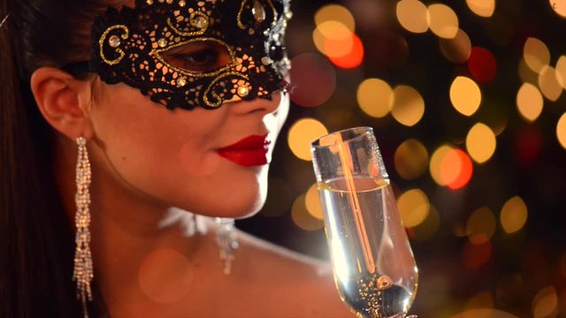 Beautiful sexy woman wearing venetian mask drinking champagne over holiday glowing background. 4K UHD video footage. Ultra high definition 3840X2160