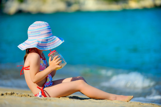 a little girl in a big beach hat is resting on the beach, drinking fresh fruit juice