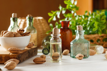 Fototapeta na wymiar Glass bottle with oil, some almond in shell around, old glass vials and greenery on a white wooden table, selective focus