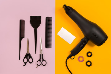 Black hair dryer, comb and scissors on pink and yellow paper background. Top view