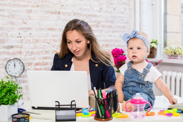 Obraz na płótnie Canvas Cheerful young beautiful businesswoman looking at laptop while sitting at her working place with her little daughter