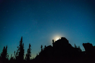 Stars and the moon against the backdrop of silhouettes of trees and rocks
