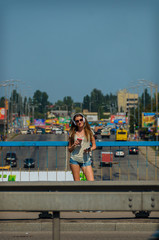 Beautiful stylish girl student stands on the car bridge with a beautiful view of the city and drinks music