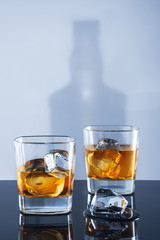Two glasses of whisky and ice on a light background with a shadow from a bottle