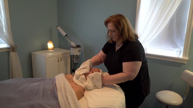 Massage therapist unwraps a hot wet towel from around a beautiful mature woman’s face as part of her facial massage. Steadicam shot.