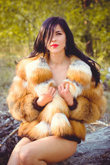 Young woman in luxurious furs with a naked body in the forest against a background of nature. Beautiful lady posing near trees in a fur coat