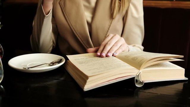Woman in a cafe drinking coffee and reading a book. Shot of woman's hand reading a book indoors. Close up. Cute girl learn and read a literature turning pages of the book holding hands closeup