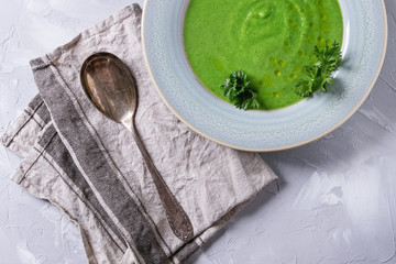 Vegetarian vegan broccoli cream soup served in blue plate with fresh parsley, broccoli, spoon, textile napkin over gray concrete background. Top view with copy space. Healthy eating.