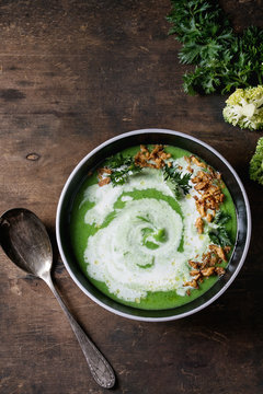 Vegetarian broccoli cream soup served in black bowl with spoon, cream, fried onion, fresh parsley and broccoli over old wooden background. Top view with space. Healthy eating.