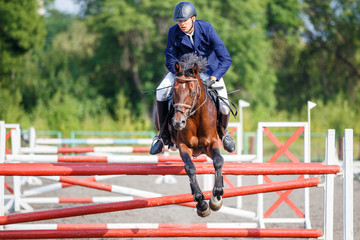 Young rider man on bay horse jumping over hurdle on show jumping competition