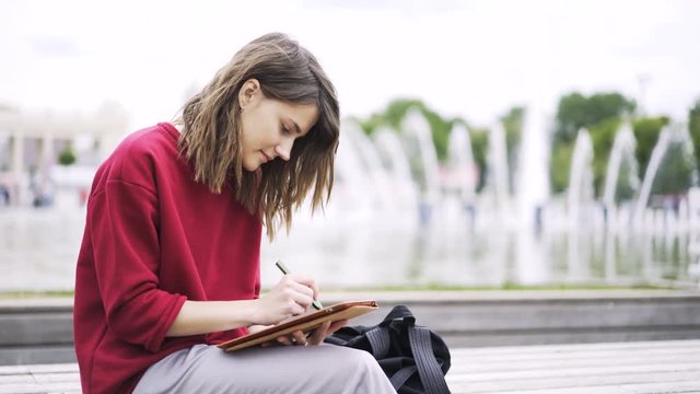 Pretty young woman with fair hair wearing a red sweater drawing at her tablet computer with a special pen while sitting near a fountain. Left to right pan real time establishing shot