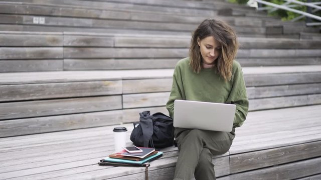 Beautiful young woman wearing a green sweatshirt sitting on the stairs outside and typing at her laptop keyboard. Locked down real time establishing shot