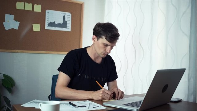 Freelancer working on laptop computer writing ideas on adhesive notes. A board with stickers and graphics at wall behind